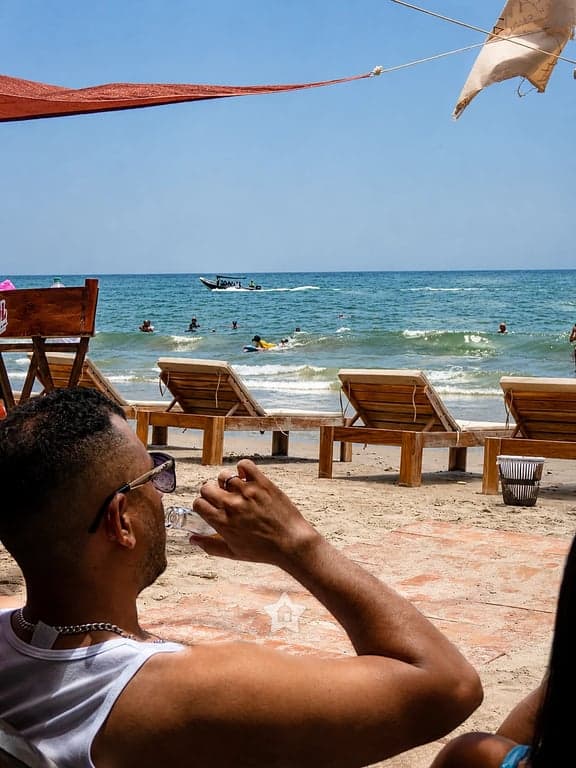 Camastros de madera frente al mar en Nakawa Beach Club, con bañistas y lanchas al fondo