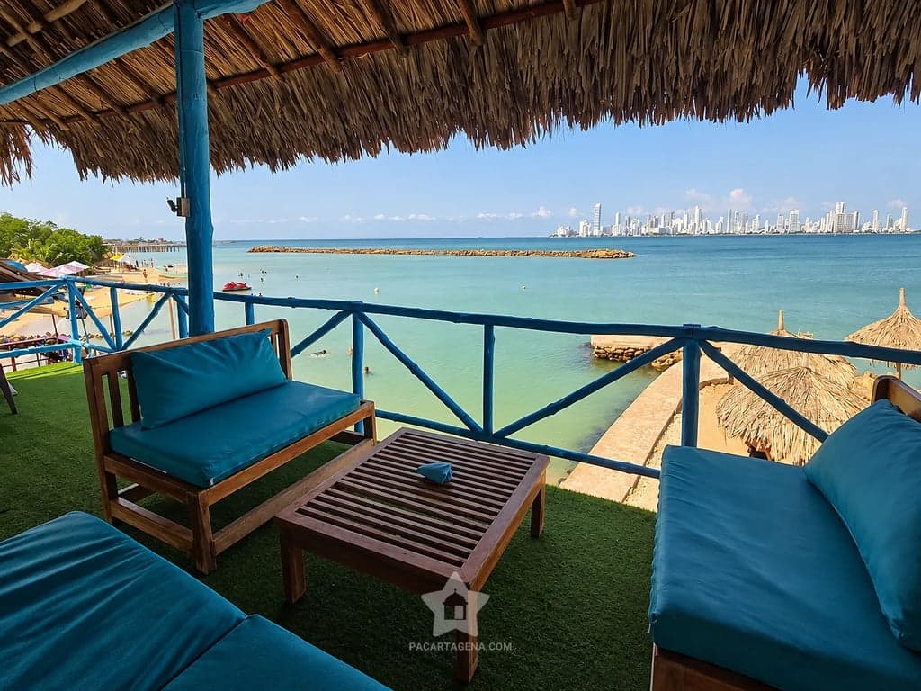 Mesa de madera entre dos sillones azules bajo techo de palma, frente a la bahía en Paraíso del Sol, Tierra Bomba.